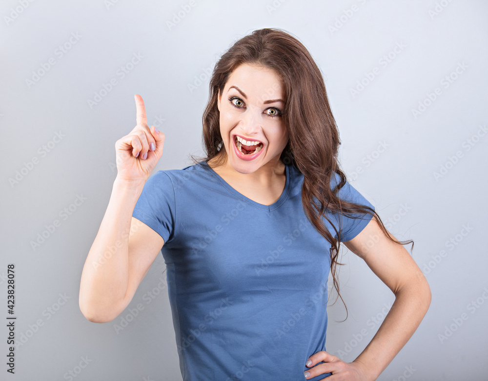 Fototapeta premium Beautiful excited casual woman with shock opened mouth pointing the finger up with smiling on empty copy space blue background. Closeup