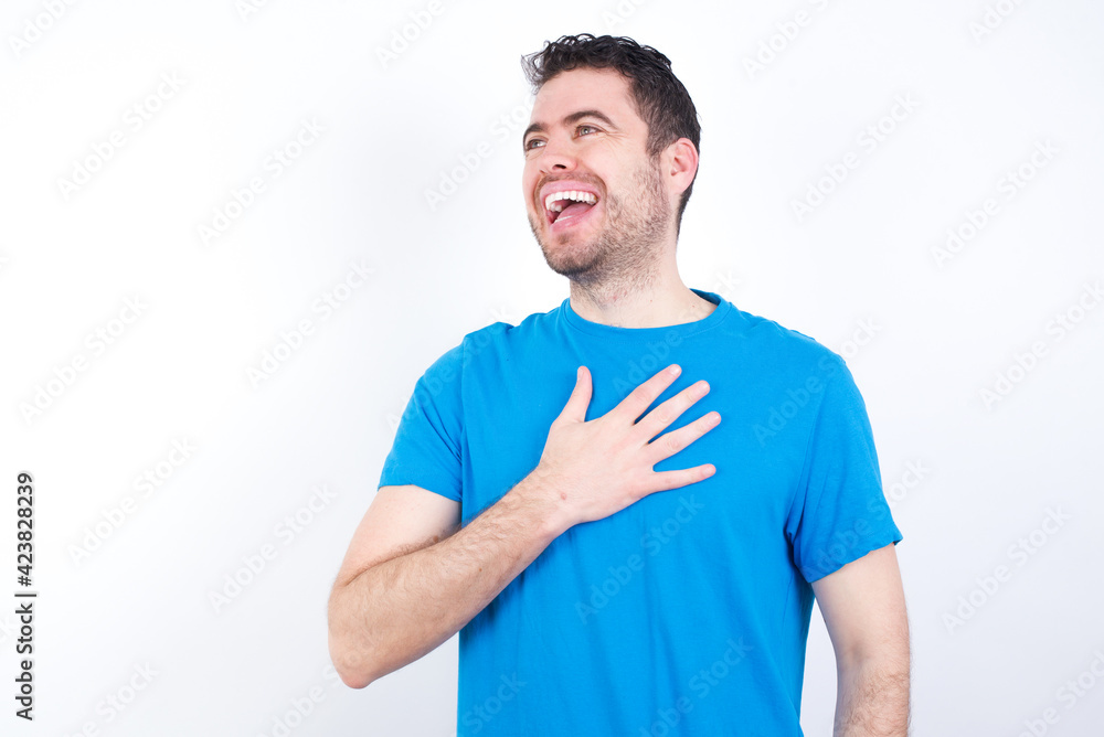 Joyful young handsome caucasian man wearing white t-shirt against white background expresses positive emotions recalls something funny keeps hand on chest and giggles happily. Joyful young handsome caucasian man wearing white t-shirt against white background expresses positive emotions recalls something funny keeps hand on chest and giggles happily.