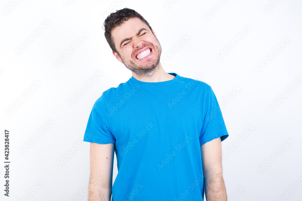 Positive young handsome caucasian man wearing white t-shirt against white background with overjoyed expression closes eyes and laughs shows white perfect teeth Positive young handsome caucasian man wearing white t-shirt against white background with overjoyed expression closes eyes and laughs shows white perfect teeth