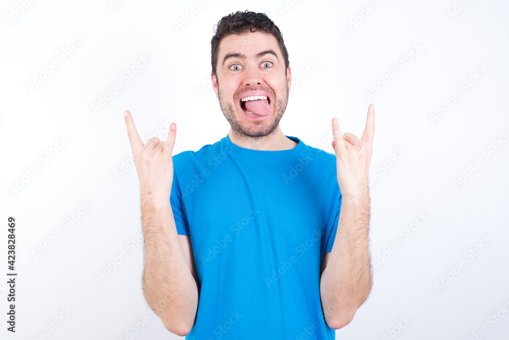 young handsome caucasian man wearing white t-shirt against white background makes rock n roll sign looks self confident and cheerful enjoys cool music at party. Body language concept. young handsome caucasian man wearing white t-shirt against white background makes rock n roll sign looks self confident and cheerful enjoys cool music at party. Body language concept.