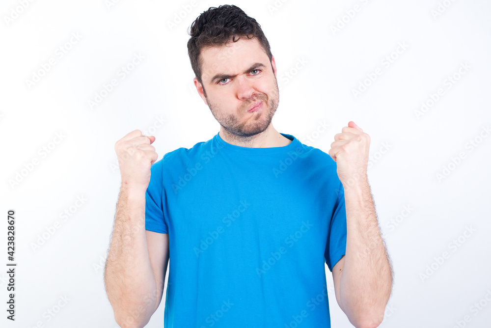 Irritated young handsome caucasian man wearing white t-shirt against white background blows cheeks with anger and raises clenched fists expresses rage and aggressive emotions. Furious model Irritated young handsome caucasian man wearing white t-shirt against white background blows cheeks with anger and raises clenched fists expresses rage and aggressive emotions. Furious model