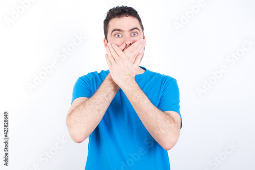 Stunned young handsome caucasian man wearing white t-shirt against white background covers both hands on mouth, afraids of something astonishing