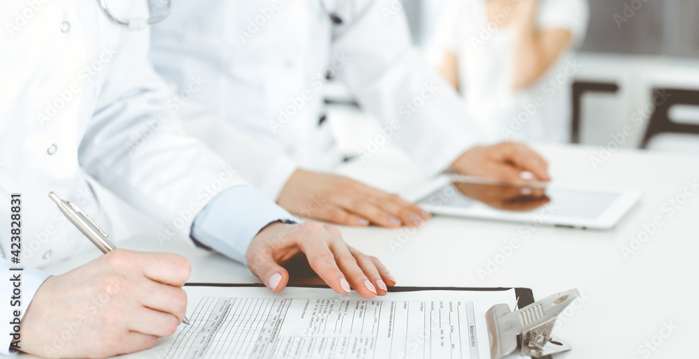 Unknown woman-doctors at work with patient at the background. Female physicians filling up medical documents or prescription while standing in hospital reception desk, close-up. Health care concept Unknown woman-doctors at work with patient at the background. Female physicians filling up medical documents or prescription while standing in hospital reception desk, close-up. Health care concept