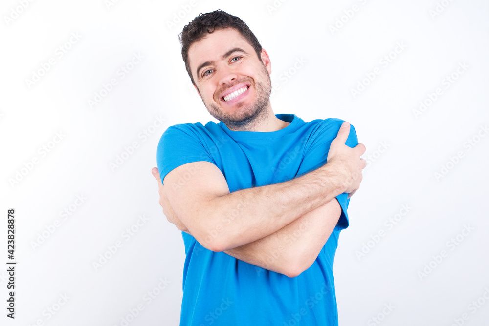 Charming pleased young handsome caucasian man wearing white t-shirt against white background embraces own body, pleasantly feels comfortable poses. Tenderness and self esteem concept Charming pleased young handsome caucasian man wearing white t-shirt against white background embraces own body, pleasantly feels comfortable poses. Tenderness and self esteem concept