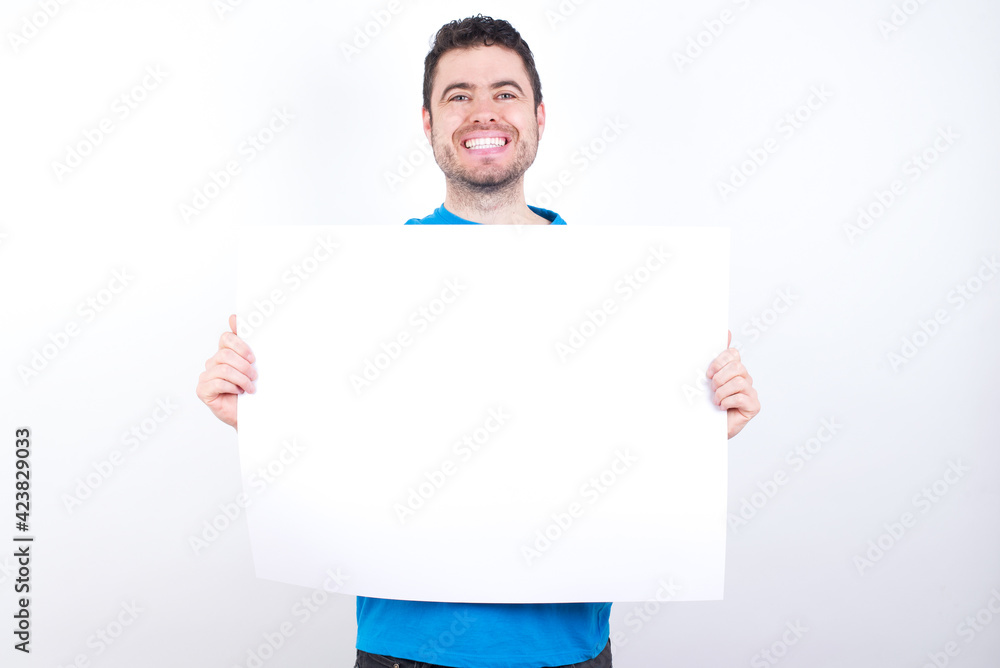 Happy young handsome caucasian man wearing white t-shirt against white background Holding Empty Paper Board Advertising Offer Text Standing. Autumn Advertisement Banner. Happy young handsome caucasian man wearing white t-shirt against white background Holding Empty Paper Board Advertising Offer Text Standing. Autumn Advertisement Banner.