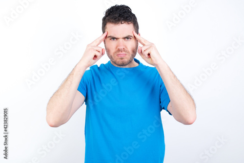 Serious concentrated young handsome caucasian man wearing white t-shirt against white keeps fingers on temples, tries to ease tension, gather with thoughts and remember important information for exam