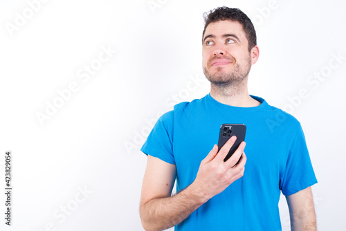 young handsome caucasian man wearing white t-shirt against white background holds mobile phone uses high speed internet and social networks has online communication. Modern technologies concept