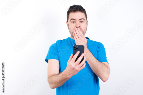 young handsome caucasian man wearing white t-shirt against white background being deeply surprised, stares at smartphone display, reads shocking news on website, Omg, its horrible!