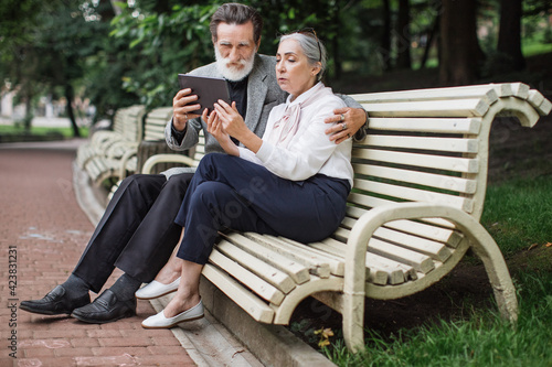 Happy mature couple using digital tablet while sitting in embrace on wooden bench. Relaxation at green park. Concept of modern technology.