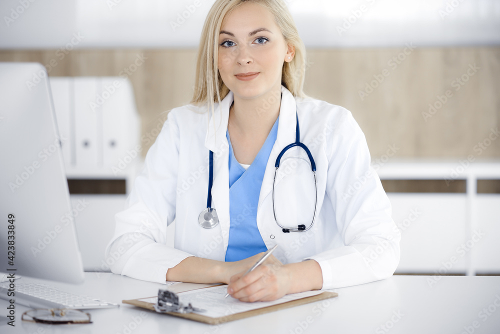 Portrate of woman-doctor at work while sitting at the desk in clinic. Blonde cheerful physician filling up medical form or prescription Portrate of woman-doctor at work while sitting at the desk in clinic. Blonde cheerful physician filling up medical form or prescription