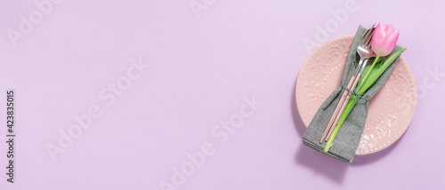 Festive table setting with pink plate, cutlery set and tulip flower on light lilac background long banner format. Festive restaurant menu template.