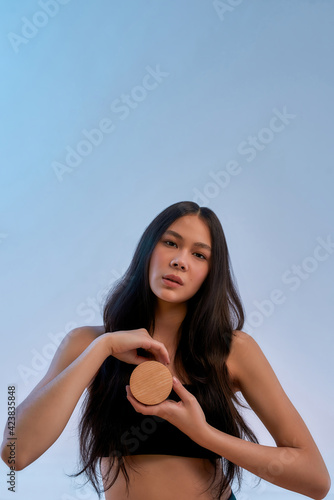 Organic facial products. Portrait of a young sensual asian woman holding beauty product and looking at camera while posing against blue background