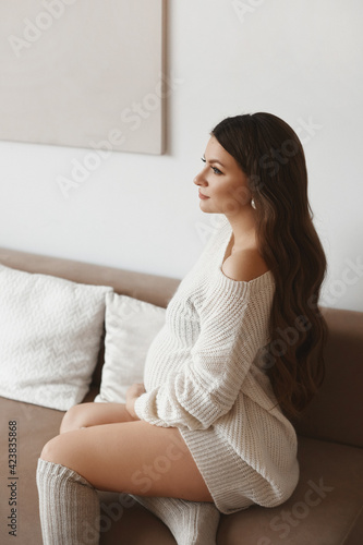 Beautiful young pregnant woman with dark hair wearing white knitted dress posing on the sofa
