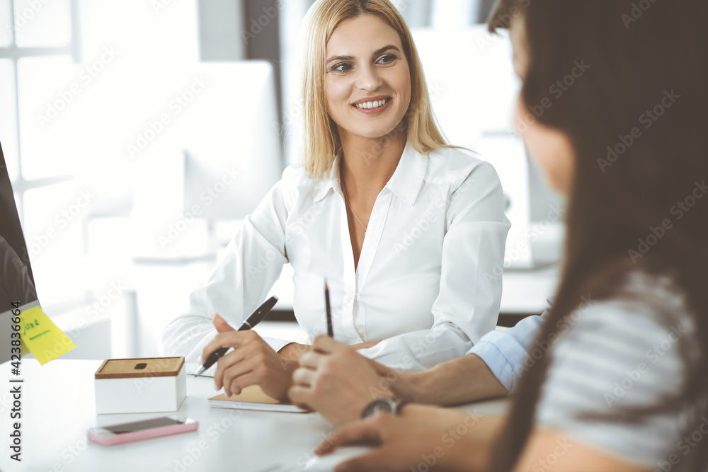 Group of business people discussing questions at meeting. Headshot of blonde businesswoman while smiling to her colleague at office negotiation. Teamwork and cooperation in corporate occupation Group of business people discussing questions at meeting. Headshot of blonde businesswoman while smiling to her colleague at office negotiation. Teamwork and cooperation in corporate occupation