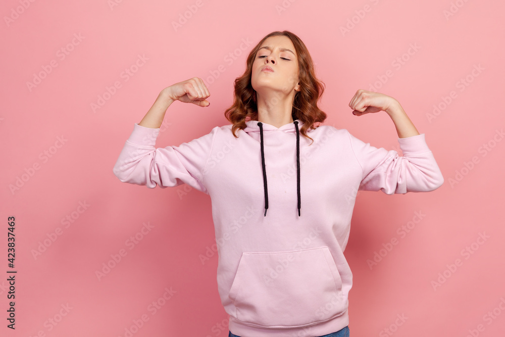 Portrait of proud young female in hoodie showing biceps, expressing power ambitions to become successful, female rights, feminism. Indoor studio shot isolated on pink background Portrait of proud young female in hoodie showing biceps, expressing power ambitions to become successful, female rights, feminism. Indoor studio shot isolated on pink background