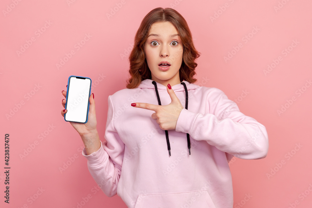 Portrait of shocked teenage curly haired girl in hoodie pointing finger on smartphone empty screen, surprised with information or discount. Indoor studio shot isolated on pink background Portrait of shocked teenage curly haired girl in hoodie pointing finger on smartphone empty screen, surprised with information or discount. Indoor studio shot isolated on pink background