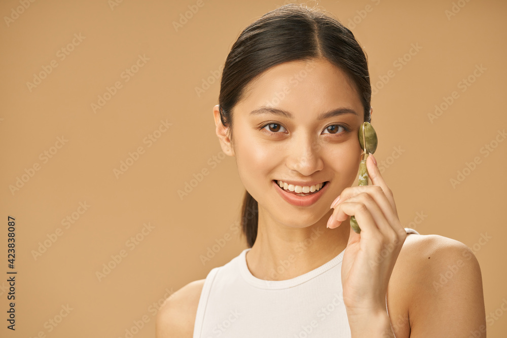 Cheerful beautiful young woman smiling at camera while using jade roller for massaging her face, posing isolated over beige background Cheerful beautiful young woman smiling at camera while using jade roller for massaging her face, posing isolated over beige background