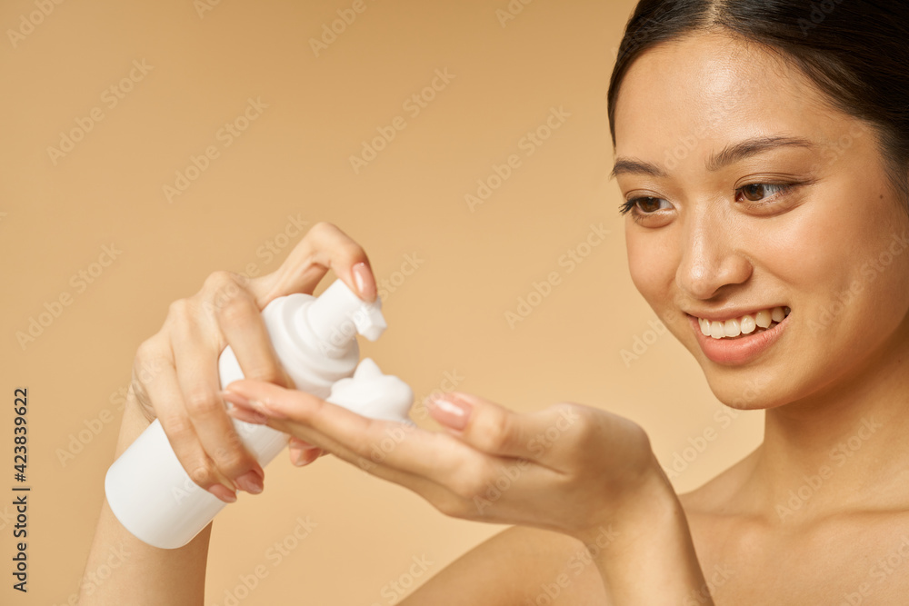 Close up portrait of cute young woman smiling, holding a bottle of gentle foam facial cleanser isolated over beige background
