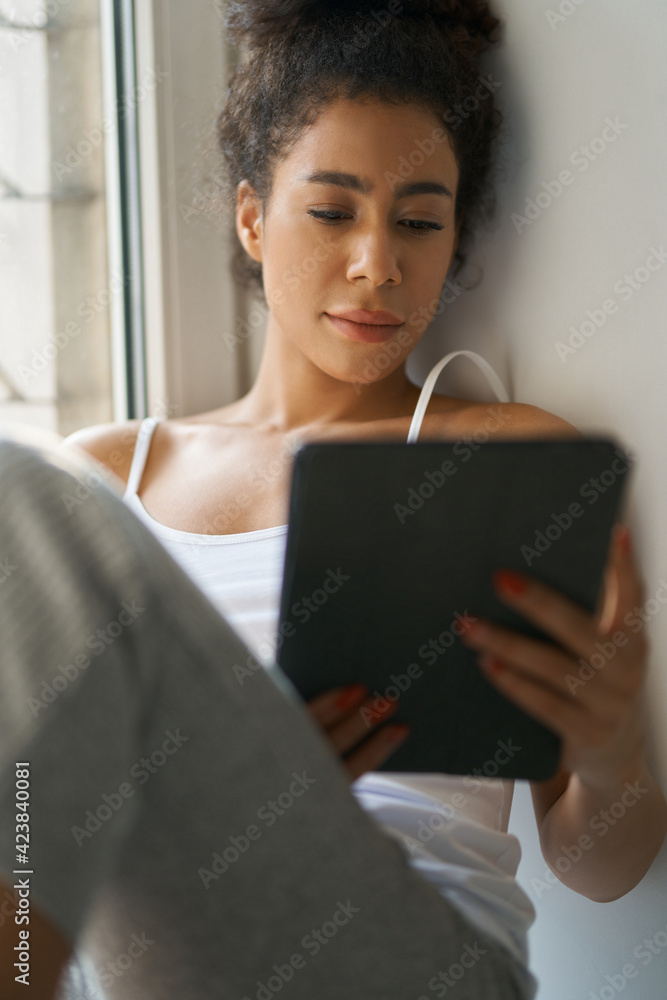Relaxed young woman reading a book using tablet pc while sitting by the window at home Relaxed young woman reading a book using tablet pc while sitting by the window at home