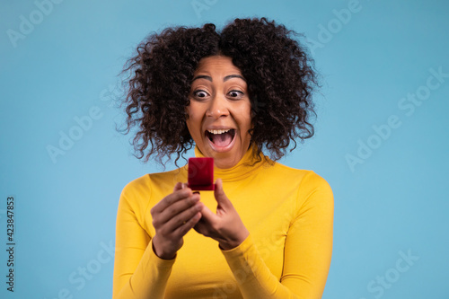 Lady screaming, jumping , she is happy to get present, proposition for marriage. Attractive african woman holding small jewelry box with proposal diamond ring on blue wall background.  © kohanova1991