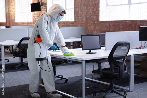 Cleaner wearing hygiene overalls, gloves and face mask disinfecting office and furniture