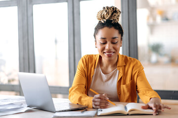 Online education. Joyful clever focused African American stylish young woman with dreadlocks, student, studying online at home, watching online video lesson, uses laptop, taking notes, smile