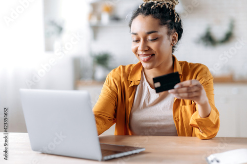 Happy young african american woman stylishly dressed, paying for online purchases, uses a laptop, holding a banking card in hand, typing credit card information on the keyboard and smiling
