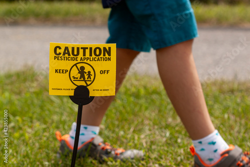A yellow yard sign warning kids and pets of the recent pesticide spraying and advices them to stay away. A kid is playing regardless. Pesticide use is a big concern.
