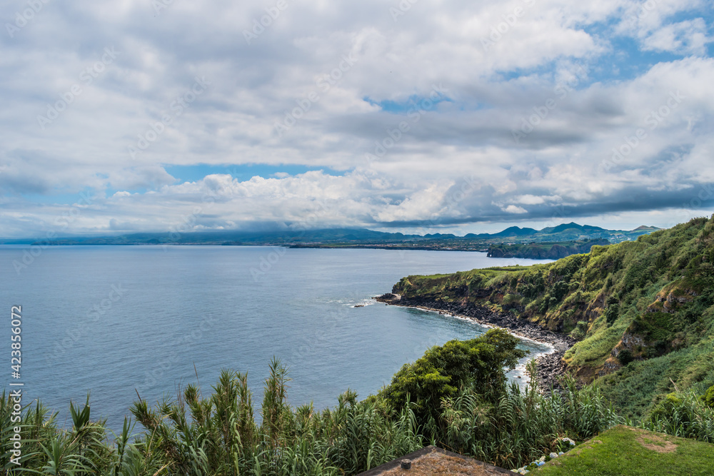Cliff and green vegetation in Fonte Grande viewpoint with gradient mountains on the horizon under cloudy sky, São Miguel - Azores PORTUGAL Cliff and green vegetation in Fonte Grande viewpoint with gradient mountains on the horizon under cloudy sky, São Miguel - Azores PORTUGAL