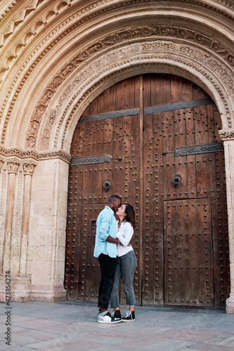 couple of young multiethnic guys kissing and laughing in front of the door of a cathedral, black guy and white girl in love © Luisphtos