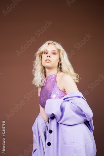 A girl in a purple jacket on a brown background in 70s, 80s, 90s style makeup, long silver earrings. The blonde is dancing, posing, fashion. A handsome woman.