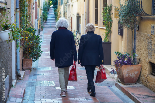 Two elegant elderly women with gray hair, red women's bags in their hands are walking up the street, a photo from the back. Antibes, France.