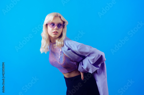 Girl in a purple jacket on a blue background in 70s, 80s, 90s style makeup, long silver earrings and purple sunglasses. Looks to the side, place for text, party.