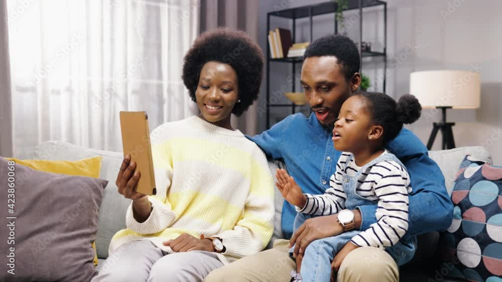 Happy young African American family sitting at home on couch with their little daughter and talking with relatives on video call waving their hands at tablet. Happy young African American family sitting at home on couch with their little daughter and talking with relatives on video call waving their hands at tablet.