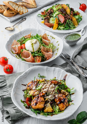 Fresh salads with grilled chicken meat, vegetables, prosciutto, mozzarella cheese, fruits, orange, greens, parmesan, lettuce, tomatoes on plates over light wooden background. Healthy food, vegan