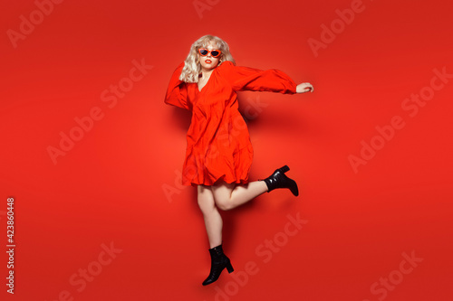 The blonde in red glasses and a dress is fashionably dressed, jumping on a red background. The woman rejoices. Model with red lips. The concept of bright style and discounts. Fashion photo.