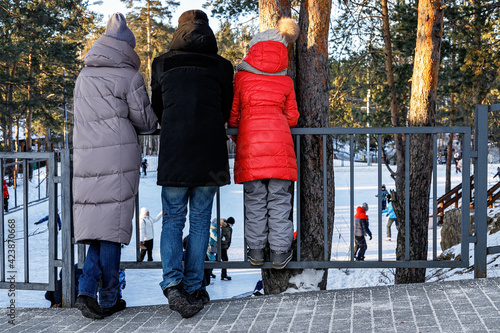 Family in bright winter clothes is watching people ice skating on a frozen lake. Fresh air, leisure activity, sunny day, forest background. Back view.