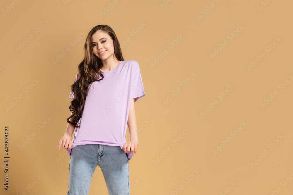 Young beautiful girl with long curly hair in a lilac T-shirt and blue jeans on a pastel orange studio background. T-shirt mockup. Young beautiful girl with long curly hair in a lilac T-shirt and blue jeans on a pastel orange studio background. T-shirt mockup.