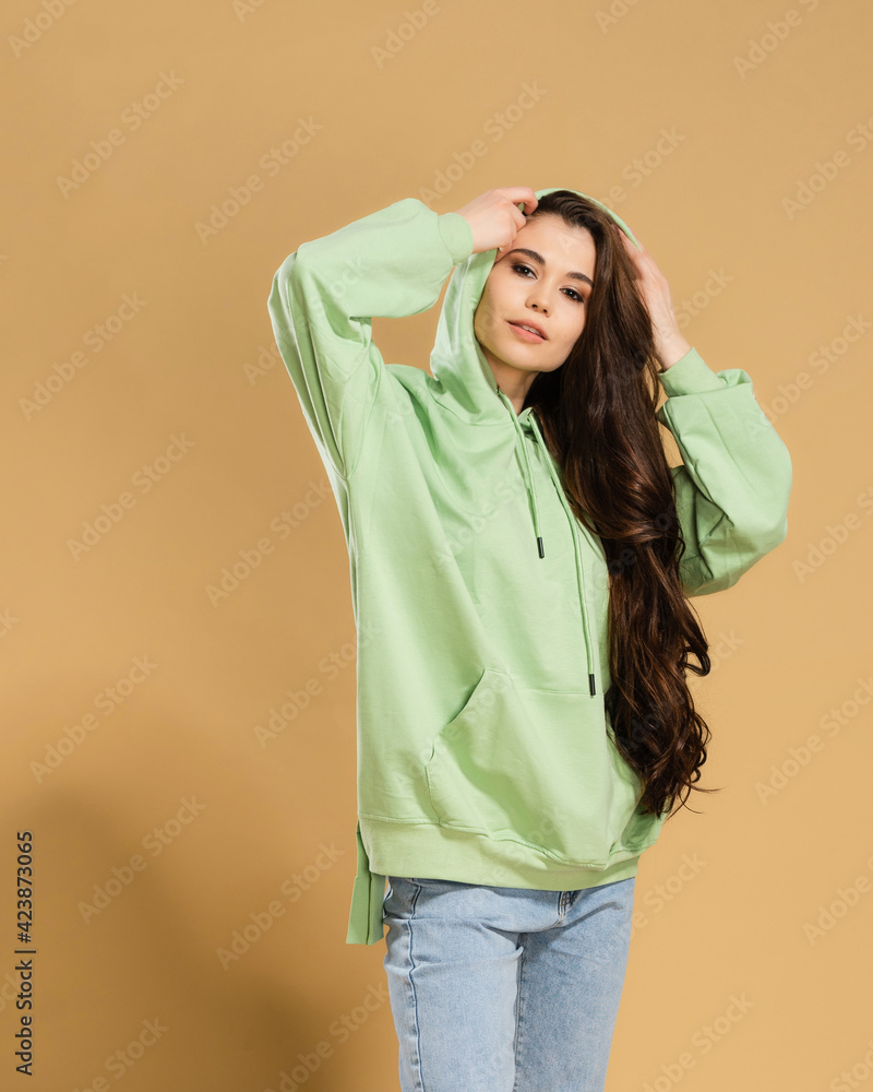 Young beautiful girl with long hair in a pistachio-colored hoodie straightens the hood on a pastel orange studio background. Young beautiful girl with long hair in a pistachio-colored hoodie straightens the hood on a pastel orange studio background.