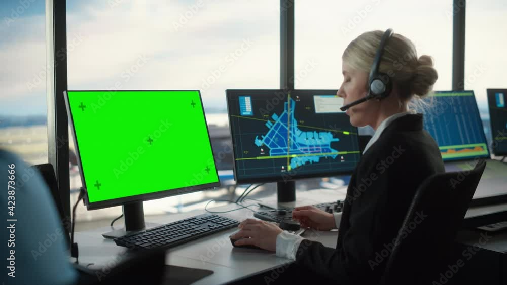 Female Air Traffic Controller with Headset Works on Computer with Green Screen Mock Up in Airport Tower. Office Room with Displays with Navigation Screens, Airplane Flight Radar Data for the Team. Female Air Traffic Controller with Headset Works on Computer with Green Screen Mock Up in Airport Tower. Office Room with Displays with Navigation Screens, Airplane Flight Radar Data for the Team.