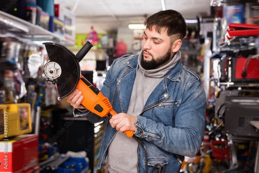 Adult cheerful positive smiling bearded man is looking on new angle grinder in tools store