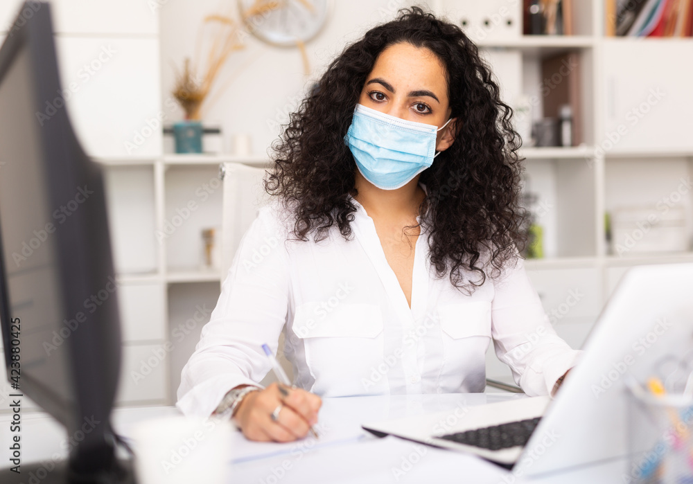 Concentrated businesswoman wearing facial mask working on laptop in business office, new normal due to coronavirus outbreak