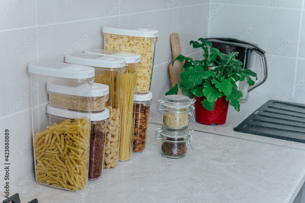 Fototapeta premium A variety of pasta,rice,cereals, nuts in containers-cans are on the kitchen table,next to a flower in a pot and a cutting board.The concept of proper convenient rational storage of food in the kitchen