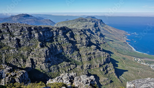 Mountain landscape at the top of Table Mountain in Cape Town. Sheer rocky slopes with green vegetation. The rugged coastline of the Atlantic Ocean is visible. Azure sky.