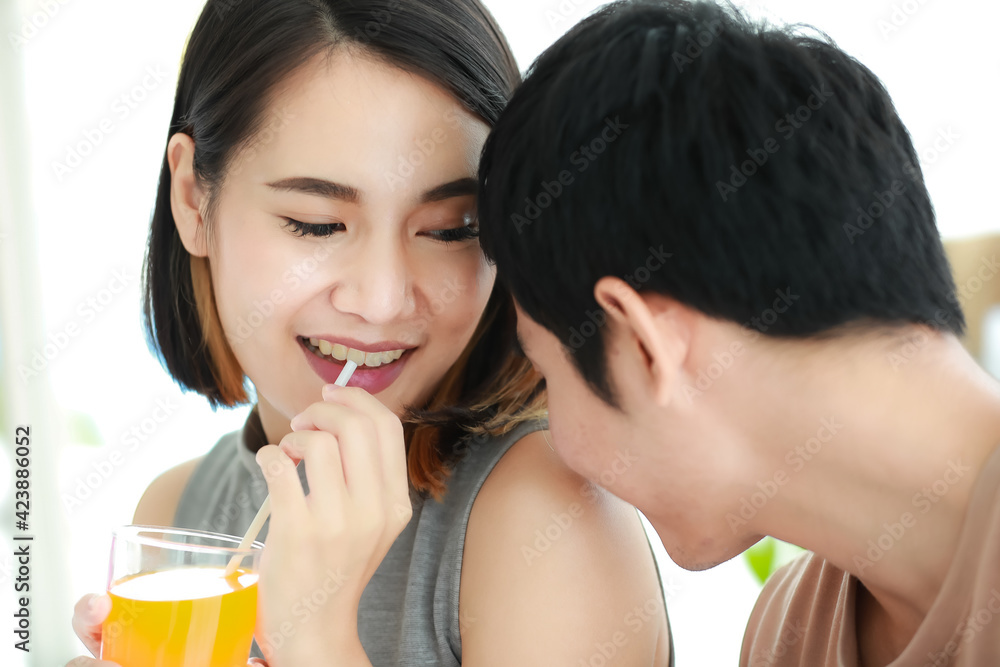 Portrait shot of adorable young Asian woman with cute smiling in casual clothes holding and drinking a glass of orange juice. Husband sitting beside his wife and kissing her shoulder with passionate Portrait shot of adorable young Asian woman with cute smiling in casual clothes holding and drinking a glass of orange juice. Husband sitting beside his wife and kissing her shoulder with passionate