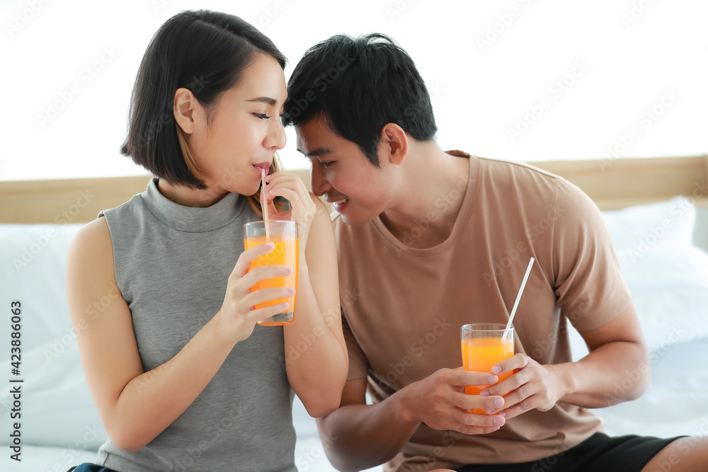 Portrait shot of adorable young Asian woman with cute smiling in casual clothes holding and drinking a glass of orange juice. Husband sitting beside his wife and kissing her shoulder with passionate Portrait shot of adorable young Asian woman with cute smiling in casual clothes holding and drinking a glass of orange juice. Husband sitting beside his wife and kissing her shoulder with passionate