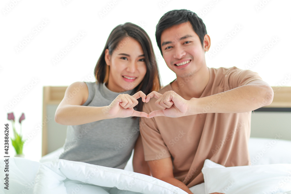 Cute smiling middle-aged Asian lover couple sitting on a bed, showing gesture in heart sign with fingers. Selective focus at the hands with a blurred face Cute smiling middle-aged Asian lover couple sitting on a bed, showing gesture in heart sign with fingers. Selective focus at the hands with a blurred face
