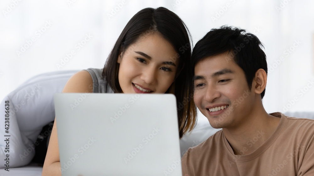 Portrait shot of cute smiling young Asian lover couple watching a movie from modern digital laptop together in the weekends. Husband playing a multiplayer online game with his wife cheering beside Portrait shot of cute smiling young Asian lover couple watching a movie from modern digital laptop together in the weekends. Husband playing a multiplayer online game with his wife cheering beside