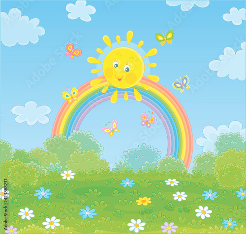 Friendly smiling sun with a colorful rainbow and butterflies merrily flittering over a green field with flowers and green bushes after warm summer rain, vector cartoon illustration