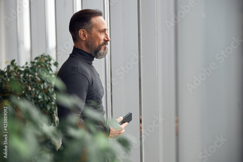 Happy smart adult caucasian businessman is standing near big windows in office space and holding smartpone